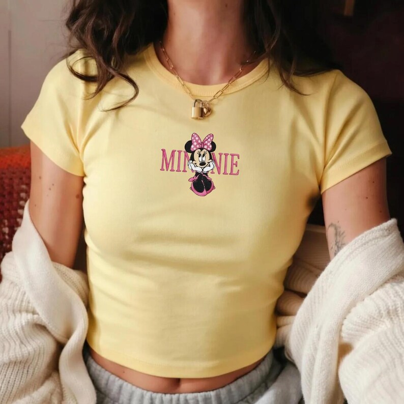 Może przedstawiać: Ż&oacute;łty top z nadrukiem Myszki Minnie. Grafika zawiera napis "Minnie" w r&oacute;żowych literach nad kresk&oacute;wkowym wizerunkiem Myszki Minnie w r&oacute;żowej kokardce i sukience.