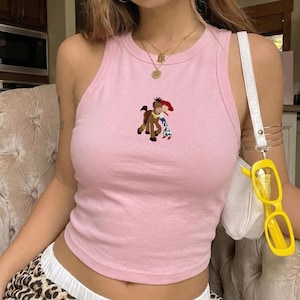 May include: A light pink tank top with a cartoon graphic of a horse and a cowgirl. The tank top has a racerback design and is made of a soft material. The tank top is a casual style.