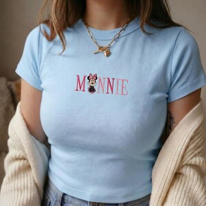 Op de afbeelding: Lichtblauw T-shirt met het geborduurde woord "MINNIE" in roze, met een Minnie Mouse-afbeelding. Het shirt heeft korte mouwen en een ronde hals. Een gouden ketting is zichtbaar. Een cr&egrave;mekleurig vest is over de schouders gedrapeerd.