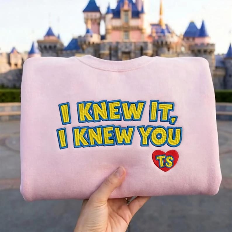 May include: A light pink crewneck sweatshirt featuring embroidered text that reads I KNEW IT, I KNEW YOU in yellow with a blue outline. Below the text is a red heart shape with the yellow letters TS embroidered in the center of the heart.