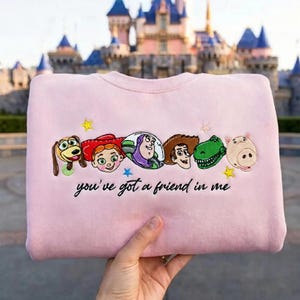 May include: A light pink crewneck sweatshirt featuring embroidered character faces from Toy Story, including Slinky Dog, Jessie, Buzz Lightyear, Woody, Rex, and Hamm, with the text "you've got a friend in me" stitched in black cursive below.