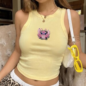 May include: A pale yellow sleeveless tank top featuring a central embroidered graphic of a pink alien character emerging from a dark hole. The top has a ribbed crew neckline and is worn with a gold necklace and a white shoulder bag with yellow glasses.