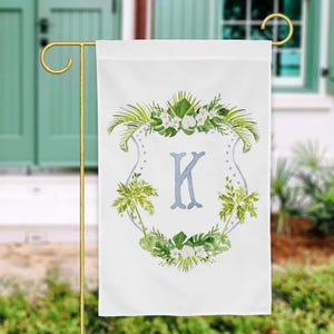 May include: White garden flag featuring a watercolor design. The flag displays a light blue letter "K" within a floral crest. Green palm fronds and white flowers surround the letter. The flag is hanging on a gold-colored metal stand.