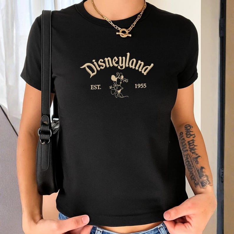 Puede incluir: Una camiseta negra de manga corta que presenta la palabra Disneyland en una fuente cursiva de color crema, con el texto EST. 1955 y una ilustraci&oacute;n de l&iacute;nea de Minnie Mouse centrada debajo del t&iacute;tulo principal en un tono crema a juego.