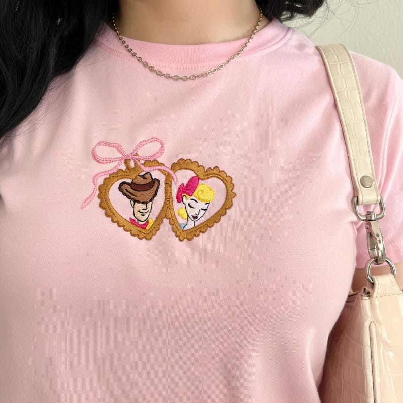 May include: A light pink t-shirt featuring an embroidered design of two cartoon characters in heart-shaped frames. The frames are gold with a scalloped edge, and the design includes a pink bow. A gold chain necklace and a cream-colored purse are also visible.