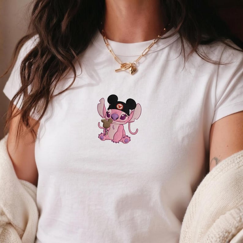 K&ouml;nnte beinhalten: Wei&szlig;es T-Shirt mit einer rosa Cartoonfigur, die einen schwarzen Hut mit Mickey Mouse-Ohren tr&auml;gt. Die Figur h&auml;lt ein gr&uuml;nes Objekt. Das Design ist auf dem T-Shirt zentriert.