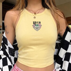 May include: A pale yellow sleeveless tank top featuring a central graphic of a green creature inside a pink heart with the black text LOVE IS printed above it. The garment has a round neckline and a fitted silhouette, worn with layered gold necklaces.