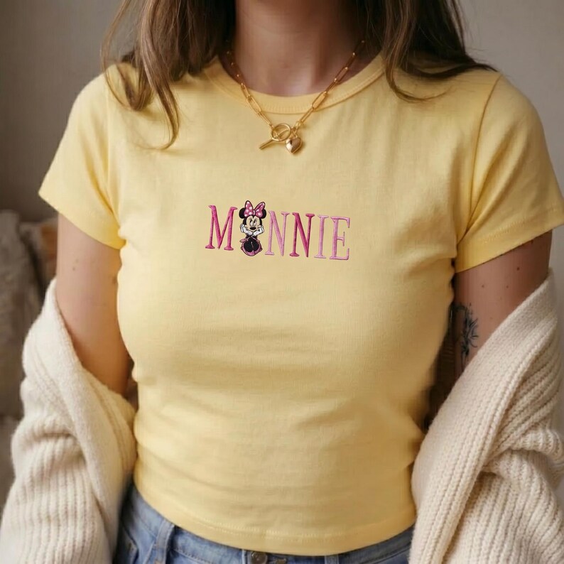 Op de afbeelding: Lichtgele t-shirt met het geborduurde woord "Minnie" in roze en een Minnie Mouse-afbeelding. Het shirt heeft korte mouwen en een ronde hals.