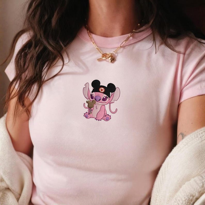 K&ouml;nnte beinhalten: Hellrosa T-Shirt mit einer Cartoonfigur mit schwarzen Mauseohren, einem schwarzen Stirnband und einem kleinen Teddyb&auml;ren. Die Figur ist rosa und lila, mit gro&szlig;en Ohren und einem niedlichen Ausdruck. Das T-Shirt hat einen runden Ausschnitt.
