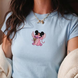K&ouml;nnte beinhalten: Hellblaues T-Shirt mit einer rosa Cartoonfigur, die einen schwarzen Hut mit Mauseohren und einem Herz tr&auml;gt. Die Figur h&auml;lt ein kleines braunes Objekt. Das Shirt ist aus weichem Material.