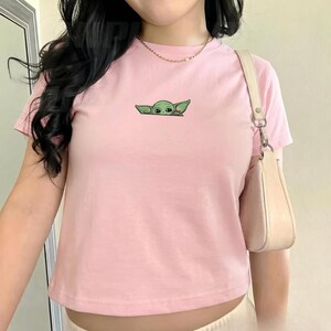 Pu&ograve; includere: Una t-shirt a maniche corte color rosa chiaro con un piccolo ricamo di una creatura verde con grandi orecchie che spunta dal centro del petto. La maglietta ha un girocollo ed &egrave; abbinata a una borsa a spalla beige con tracolla.