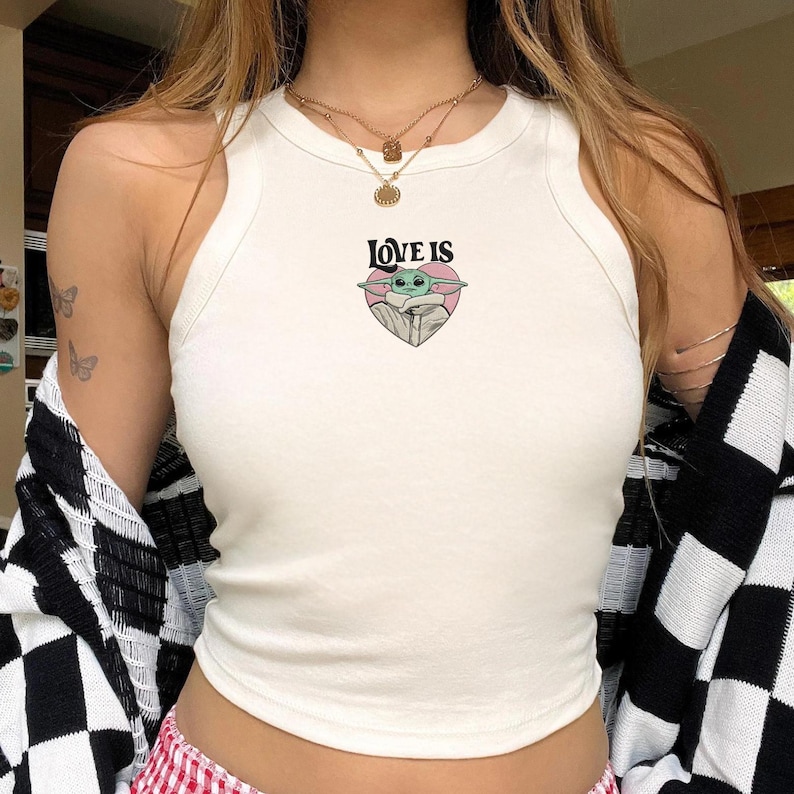 May include: A white sleeveless ribbed tank top featuring a central graphic of a green alien character inside a pink heart with the text LOVE IS printed in black above the heart. The top has a high crew neckline and a fitted silhouette.