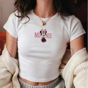 Może przedstawiać: Biały top z grafiką Minnie Mouse. Grafika zawiera słowo "MINNIE" w r&oacute;żowych literach nad kresk&oacute;wkowym wizerunkiem Minnie Mouse w r&oacute;żowej kokardce i sukience. Koszulka ma kr&oacute;tkie rękawy.