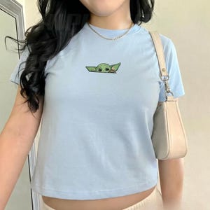 Pu&ograve; includere: Una t-shirt a maniche corte color azzurro con un piccolo ricamo di una creatura verde dalle grandi orecchie che spunta al centro del petto. La maglietta &egrave; abbinata a una borsa a spalla beige con una finitura strutturata e liscia.