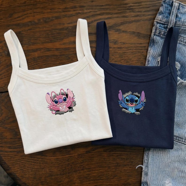 May include: A white tank top featuring an embroidered pink alien character and a navy blue tank top featuring an embroidered blue alien character. Both tops display the characters emerging from a torn fabric design on the center of the chest.