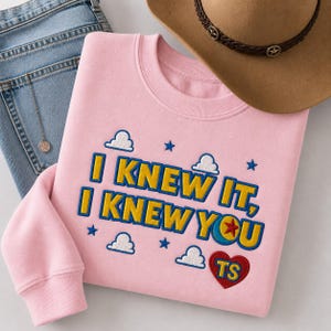 May include: A light pink crewneck sweatshirt featuring embroidered text that reads I KNEW IT, I KNEW YOU in yellow with blue outlines. The design includes white cloud icons, blue stars, a red heart with yellow TS letters, and a blue and yellow ball icon.