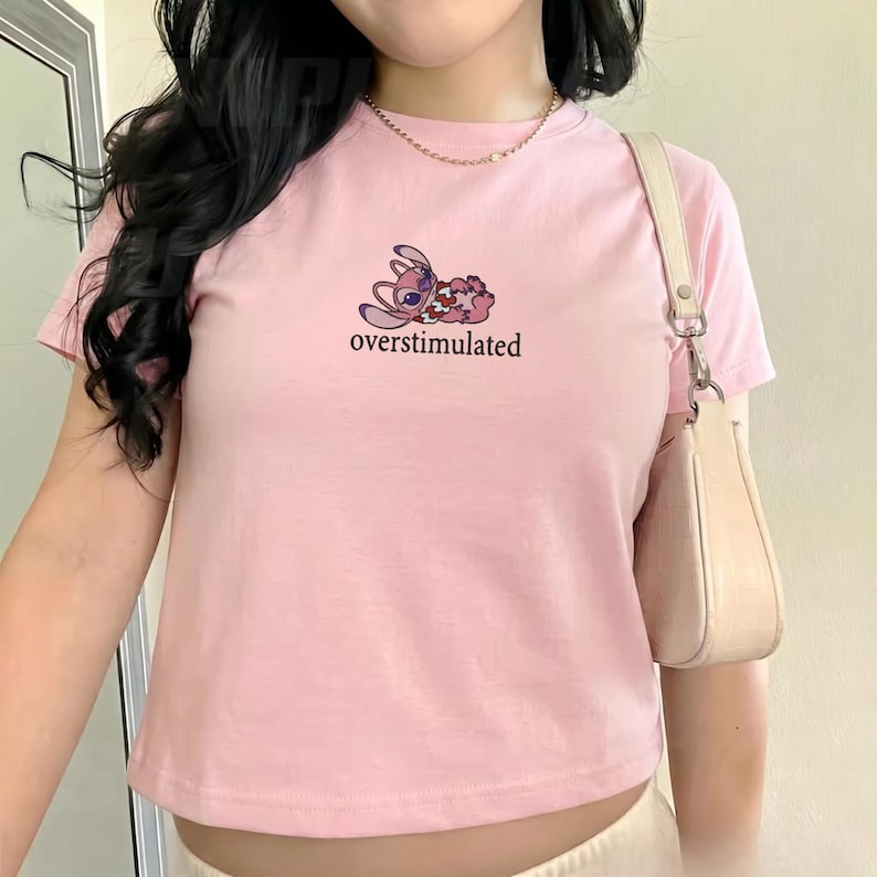 May include: Pink t-shirt with a cartoon graphic of a character and the word "overstimulated" in a black font. The shirt is short-sleeved and has a crew neck. A small, light-colored purse hangs from the shoulder.