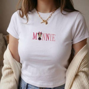Op de afbeelding: Wit T-shirt met het geborduurde woord "MINNIE" in roze, met een Minnie Mouse-afbeelding. Het shirt heeft een ronde hals en korte mouwen. Een gouden ketting met een hartvormige hanger is zichtbaar.