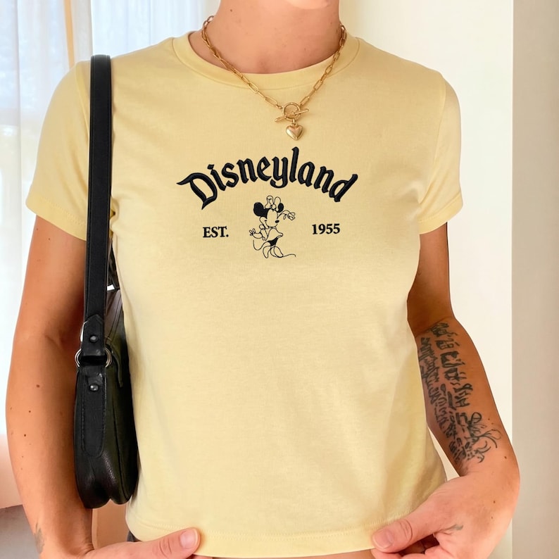 Puede incluir: Una camiseta de manga corta de color amarillo p&aacute;lido con el texto negro Disneyland, EST. 1955 y un gr&aacute;fico de Minnie Mouse. La camiseta tiene cuello redondo y se lleva con un collar de cadena dorada con colgante de coraz&oacute;n y un bolso negro.