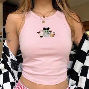 May include: A light pink sleeveless tank top featuring a central embroidered graphic of a green character with large ears wearing black mouse ears, holding a drink, and surrounded by various snacks including a cupcake, a donut, and an ice cream cone.