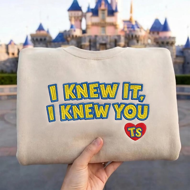 May include: A beige crewneck sweatshirt featuring embroidered text that reads I KNEW IT, I KNEW YOU in yellow with blue outlines. Below the text is a red heart shape containing the yellow embroidered letters TS with a blue outline.