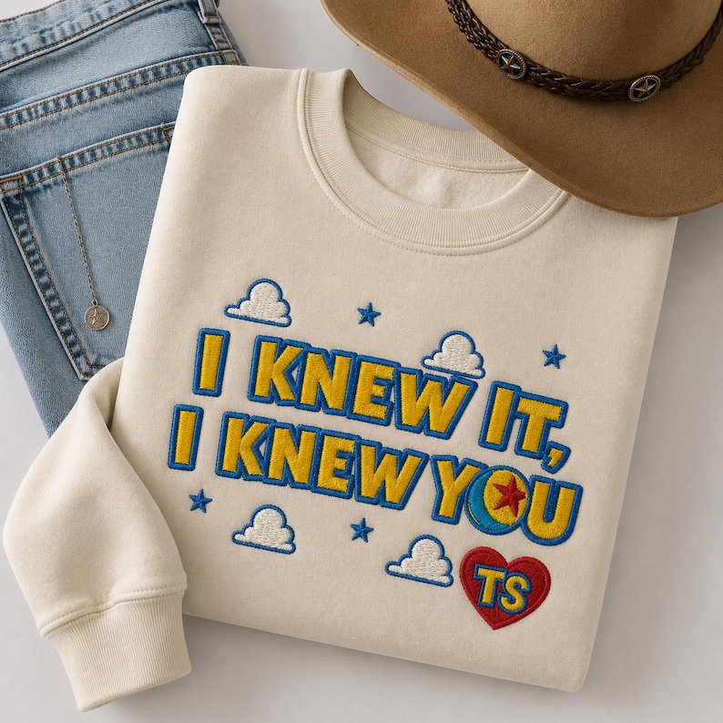 May include: A cream-colored crewneck sweatshirt featuring embroidered text that reads I KNEW IT, I KNEW YOU in yellow with blue outlines. The design includes small white clouds, blue stars, and a red heart containing the yellow letters TS.