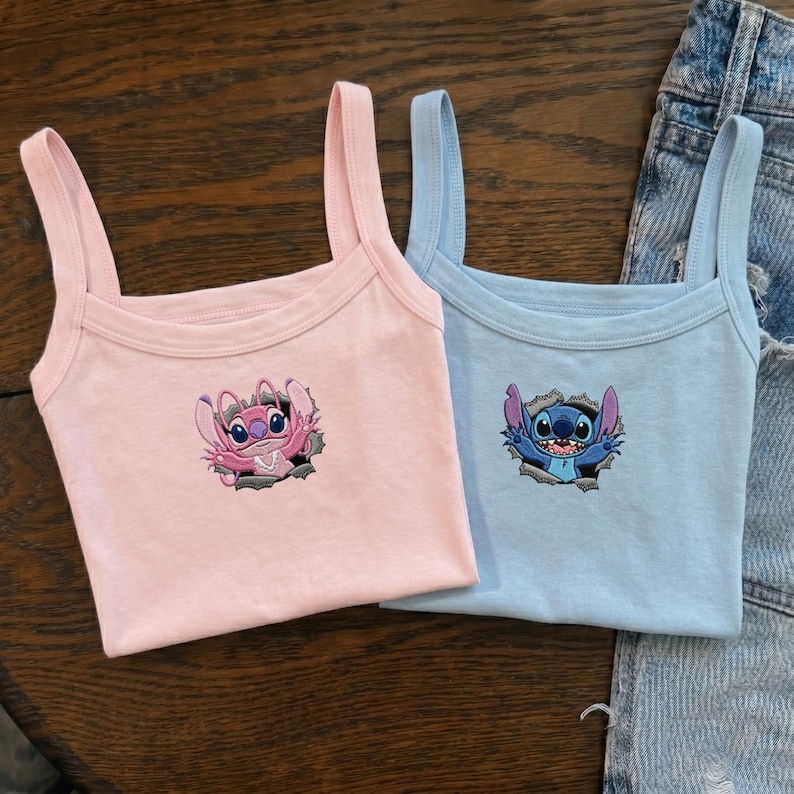 May include: A light pink tank top featuring an embroidered pink alien character and a light blue tank top featuring an embroidered blue alien character, both depicted as if emerging from a torn hole in the fabric, with wide shoulder straps.