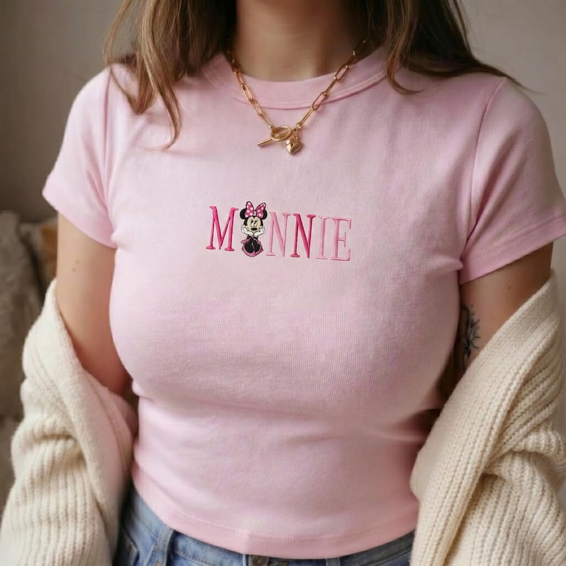 Op de afbeelding: Lichtroze T-shirt met het geborduurde woord "MINNIE" in een donkerdere roze lettertype. De letter "O" is vervangen door een Minnie Mouse afbeelding. Het shirt heeft korte mouwen en een ronde hals.