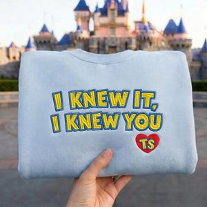 May include: A light blue crewneck sweatshirt featuring embroidered yellow text with a blue outline that reads I KNEW IT, I KNEW YOU. Below the text is a red heart shape with the yellow embroidered letters TS inside the heart.