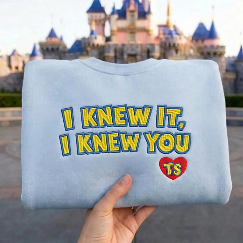 May include: A light blue crewneck sweatshirt featuring embroidered yellow text with a blue outline that reads I KNEW IT, I KNEW YOU. Below the text is a red heart shape with the yellow embroidered letters TS inside the heart.