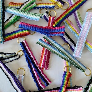 Può includere: Una collezione di portachiavi in ​​nastro tessuto colorato con anelli dorati. I nastri sono in vari motivi e colori, tra cui arcobaleno, rosa, blu, verde e bianco.