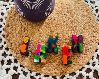 Puede incluir: Letras de crayones de colores que deletrean la palabra "LAKE". Las letras están hechas de crayones derretidos en varios colores, incluyendo rosa, naranja, amarillo y azul. Las letras están dispuestas sobre una estera tejida con flecos blancos.