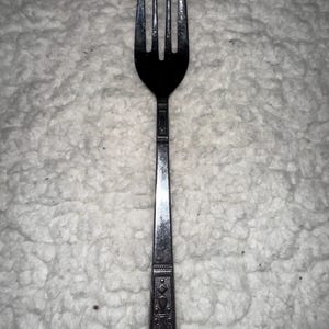Puede incluir: Un tenedor plateado con cuatro púas y un mango decorativo. El mango presenta un patrón detallado de diamantes y formas florales. El tenedor está hecho de un metal brillante y reflectante.