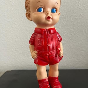 Peut inclure: Une poupée en caoutchouc vintage avec un chapeau bleu, une chemise et un short rouges et des chaussures rouges. La poupée a un visage souriant avec des yeux bleus et une bouche rouge.