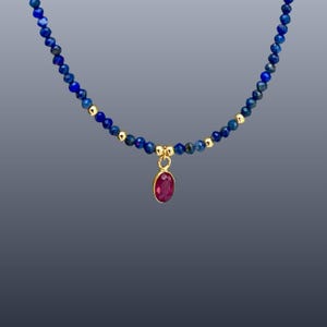 May include: A beaded necklace featuring small, faceted deep blue lapis lazuli stones interspersed with tiny gold-toned metallic beads. A single oval-shaped, faceted dark pink ruby-colored gemstone pendant hangs from a gold-toned metal bail.