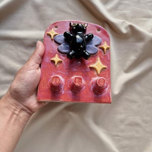 May include: A red ceramic wall hook featuring a black cat figure, purple wings, and yellow star accents. The hook has three pegs for hanging items. The cat has a glossy finish.