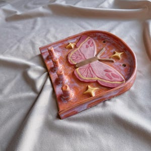May include: A ceramic butterfly-shaped trinket dish in shades of orange, pink, and yellow. The dish features a raised butterfly design with yellow accents and several small star-shaped embellishments. It has four small, cylindrical posts.