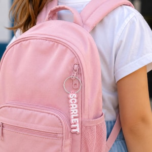 May include: A solid light pink backpack featuring a main zippered compartment, a smaller front zippered pocket, and a side mesh pocket. A silver circular keychain with the name SCARLETT in white letters on a pink background hangs from the zipper.