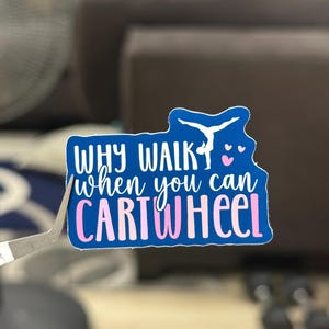 Può includere: Un adesivo blu con la scritta "WHY WALK when you can CARTWHEEL" in bianco e rosa. L'adesivo presenta una silhouette di ginnasta e piccoli cuori rosa. L'adesivo è tenuto da una pinzetta.