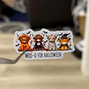 May include: A sticker featuring four Highland cows dressed in Halloween costumes: a pumpkin, a vampire, a mummy, and a witch. The text "MOO-D FOR HALLOWEEN" is below the cows. The sticker is held by tweezers.