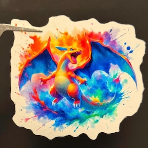 May include: A watercolor illustration of a fiery orange and yellow Pokemon character with blue wings and a tail. The character is surrounded by colorful splashes of paint.
