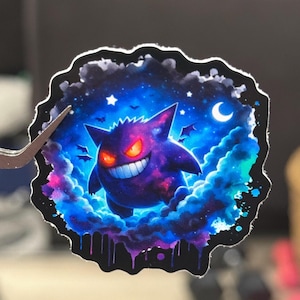 Puede incluir: Una pegatina con un personaje de dibujos animados morado con una amplia sonrisa y ojos rojos brillantes, sobre un fondo cósmico de tonos azules, morados y rosas. El personaje está rodeado de nubes estilizadas, estrellas y una luna creciente.