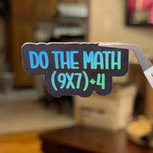 May include: A black sticker with the text "DO THE MATH (9X7)+4" in blue and green gradient lettering. The sticker is held by a pair of silver tweezers, with a blurred background.