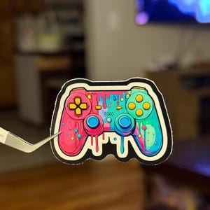 May include: A colorful sticker of a video game controller with dripping paint. The controller is pink, blue, green, and yellow.