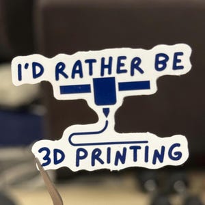 Puede incluir: Una pegatina blanca con texto azul y un gráfico de impresora 3D. El texto dice "I'D RATHER BE 3D PRINTING". La pegatina está sujeta con unas pinzas, lo que sugiere que es un artículo pequeño, posiblemente para un portátil o una botella de agua.
