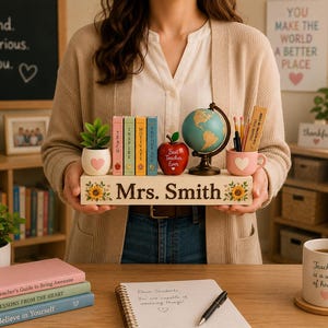 May include: A wooden desk sign with the name Mrs. Smith, decorated with sunflowers. It holds a small potted plant with a heart, four miniature books labeled Teach, Inspire, Motivate, Encourage, a red apple ornament, a globe, and a pencil cup.