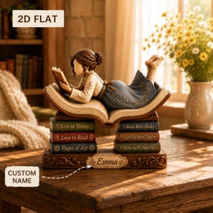 Può includere: Una statuetta decorativa di una persona sdraiata su un libro aperto, poggiato su due pile di tre libri ciascuna. I libri riportano titoli come Lost in Stories, Love to Read, Pages of Joy, Read More Books, Just One More Chapter e Books Are My Happy Place. Una base mostra il nome personalizzato Emma.