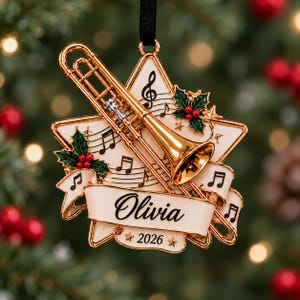 Pode incluir: Um enfeite de Natal em forma de estrela com um trombone dourado, notas musicais numa pauta e folhas de azevinho com bagas vermelhas. Uma faixa cor de creme exibe o nome Olivia e o ano 2026 em letras pretas.
