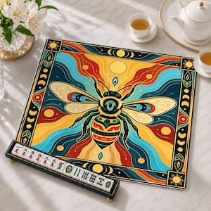Può includere: Un tappetino da gioco quadrato con un'illustrazione centrale di un'ape su motivi vorticosi gialli, arancioni e blu, incorniciato da soli e fasi lunari. Un supporto nero contiene una fila di tessere bianche del Mahjong con caratteri.