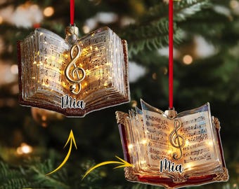 Pode incluir: Duas decorações de Natal em forma de livros abertos, com notas musicais e uma clave de sol. Os ornamentos têm uma clave de sol dourada e o nome "Mia" na parte inferior. O texto "2D FLAT NOT 3D" é visível no canto superior direito.
